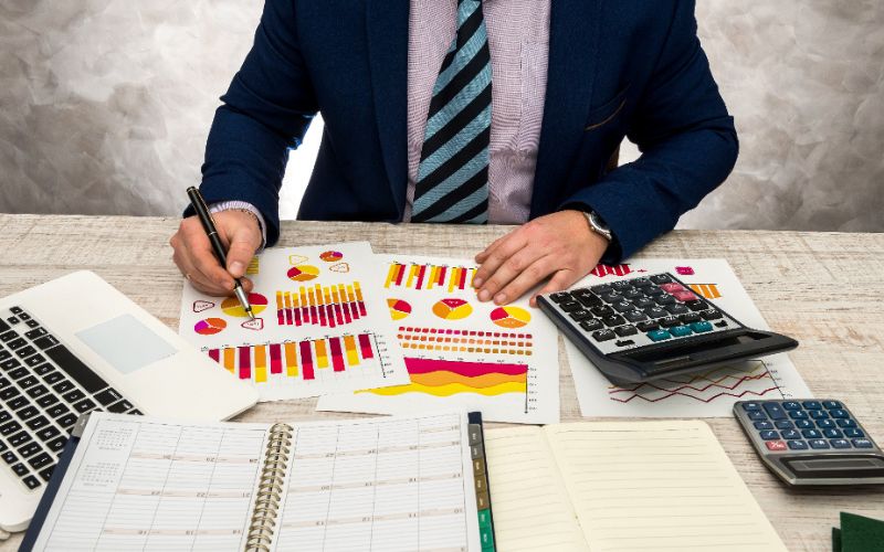 A professional man in a suit and tie works at a desk, using a laptop and reviewing papers for a tally course.