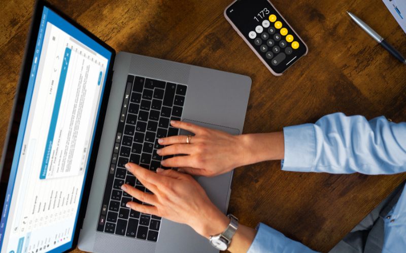 A person typing on a laptop computer with a calculator on the side.