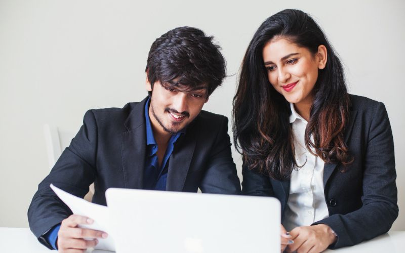 A man and woman in business attire sitting at a desk, focused on a laptop screen.